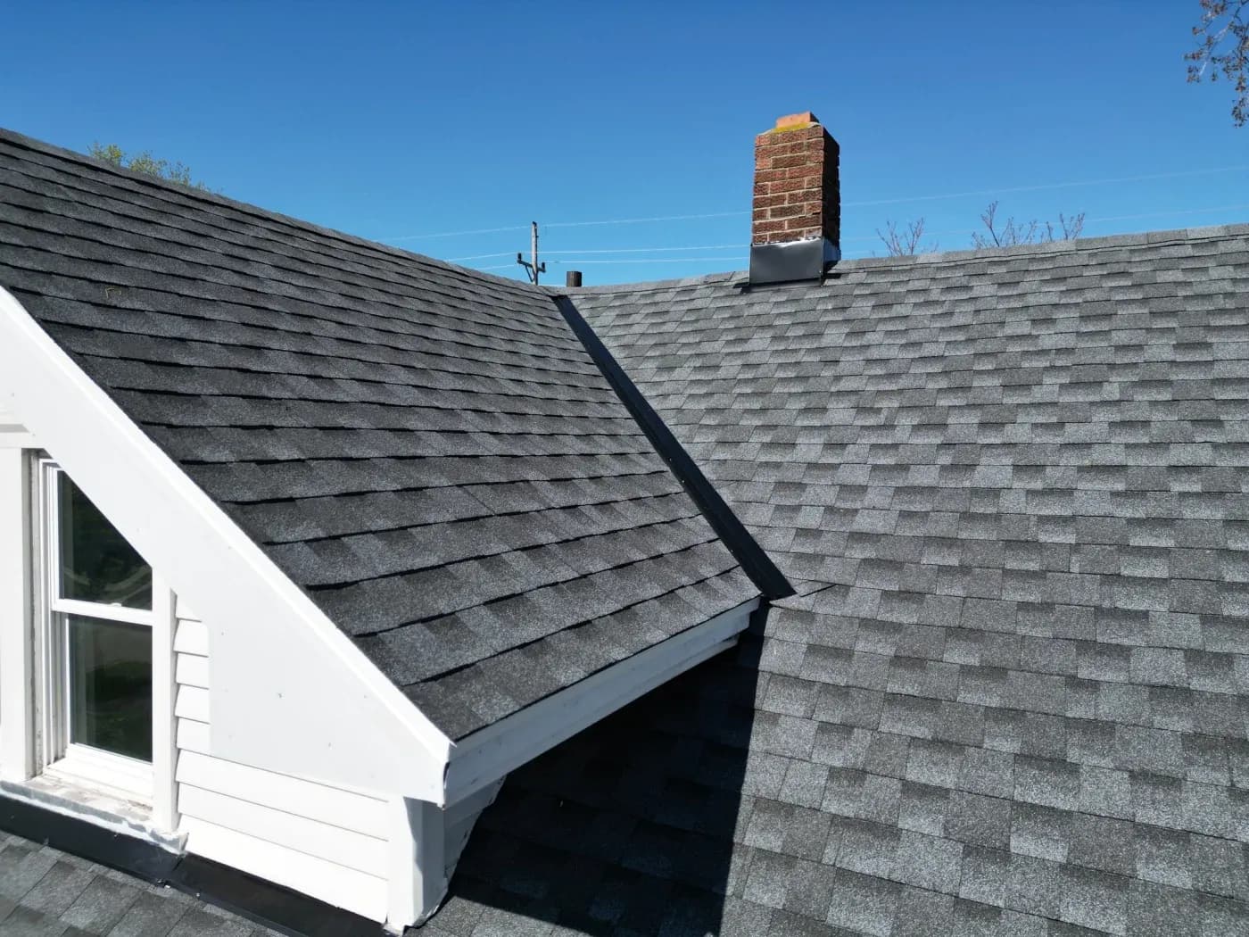 Newly installed gray asphalt shingle roof with brick chimney, metal flashing, and clear blue sky in background