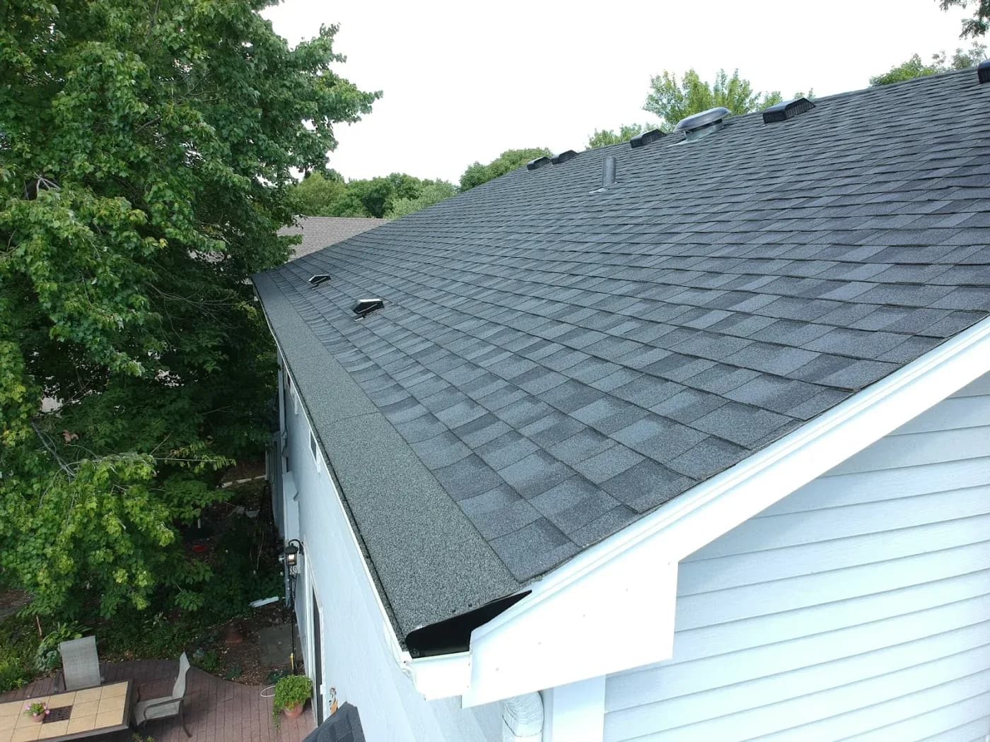 Newly installed dark gray asphalt shingle roof on a house with white gutters and siding, surrounded by green trees