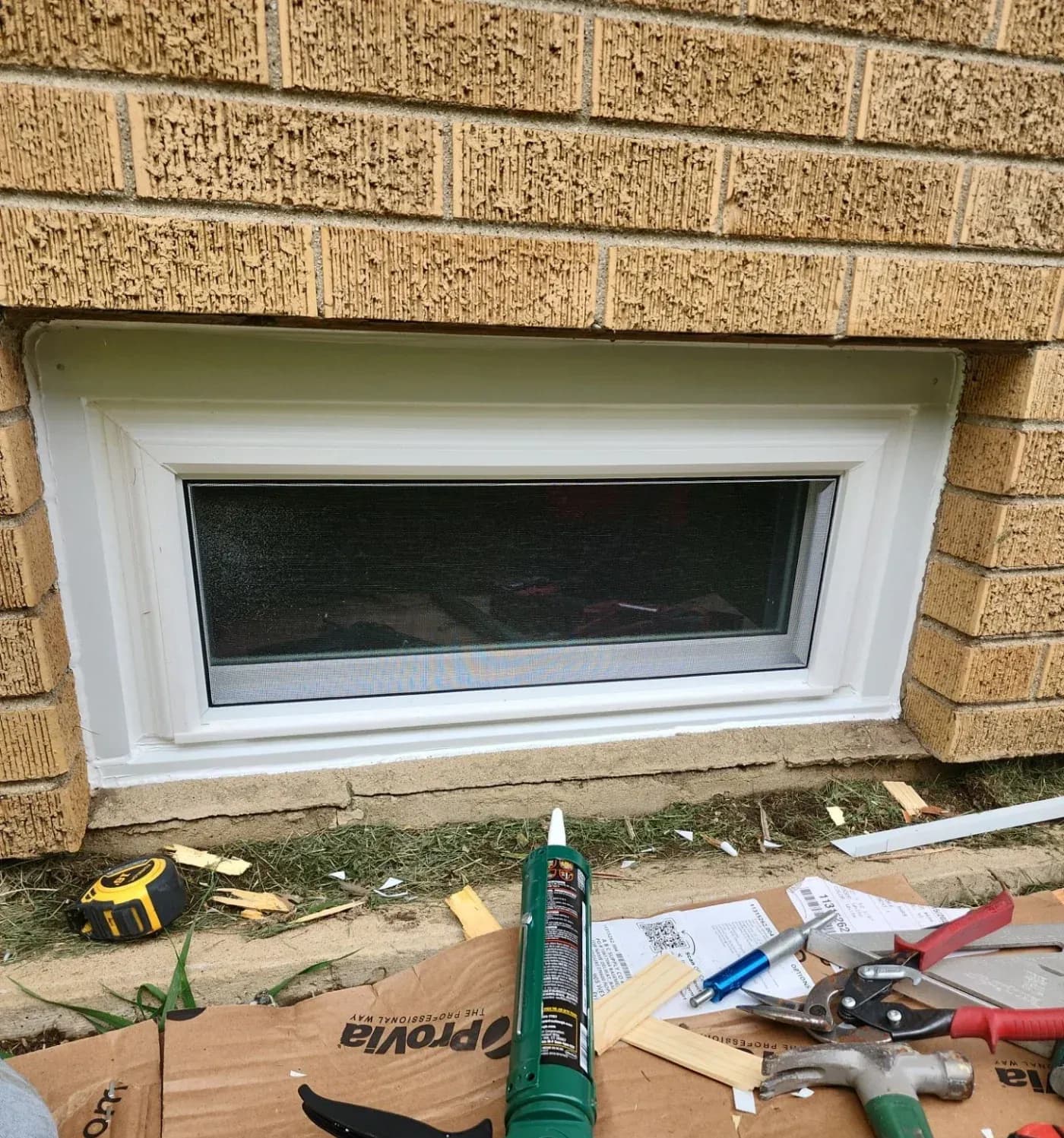 Basement window with white frame and dark screen installed in tan brick wall, surrounded by construction materials and tools including caulk, tape measure, and pliers