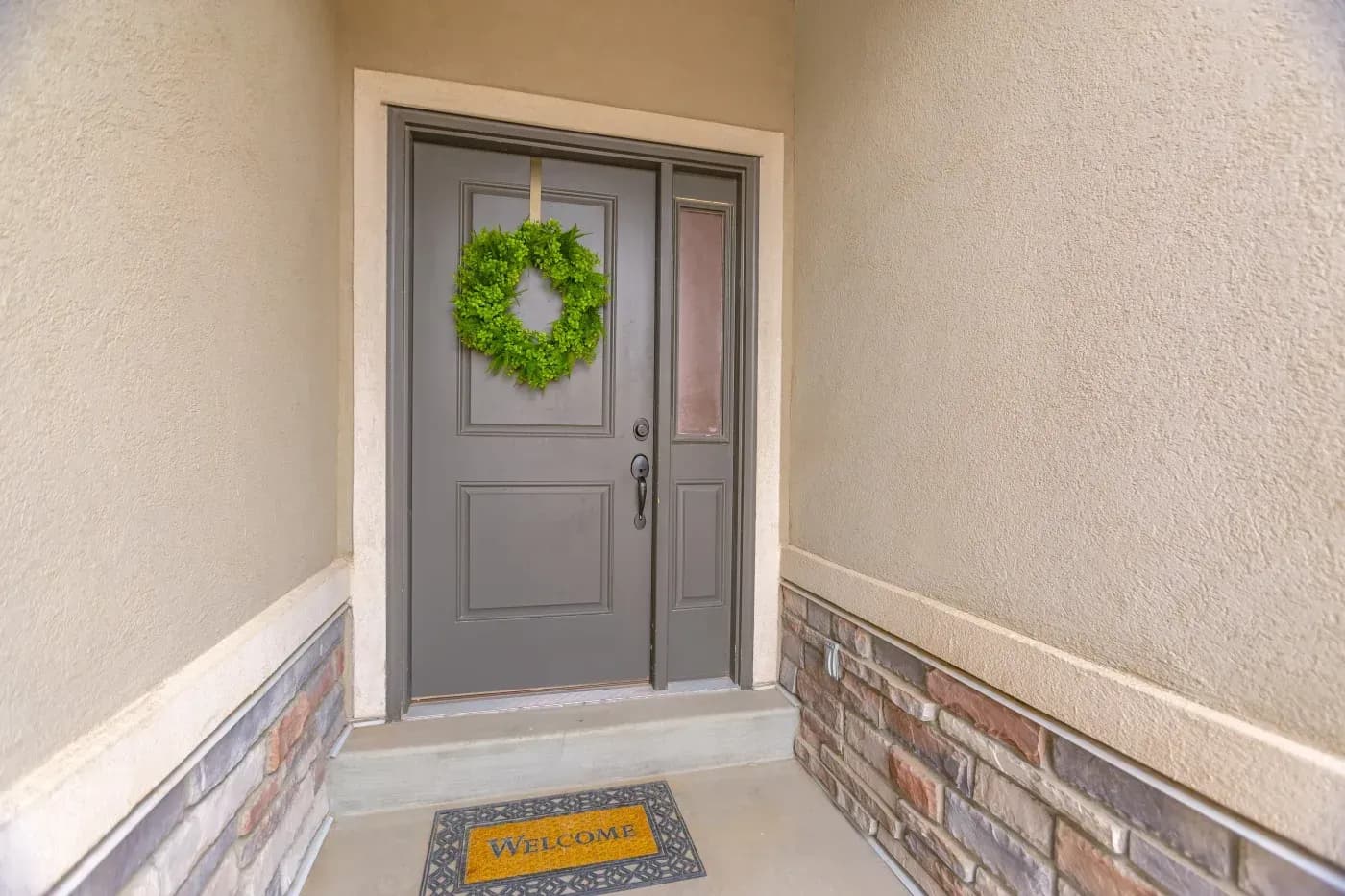 Gray front door with green wreath hanging above, sidelite window, and welcome mat on porch entrance