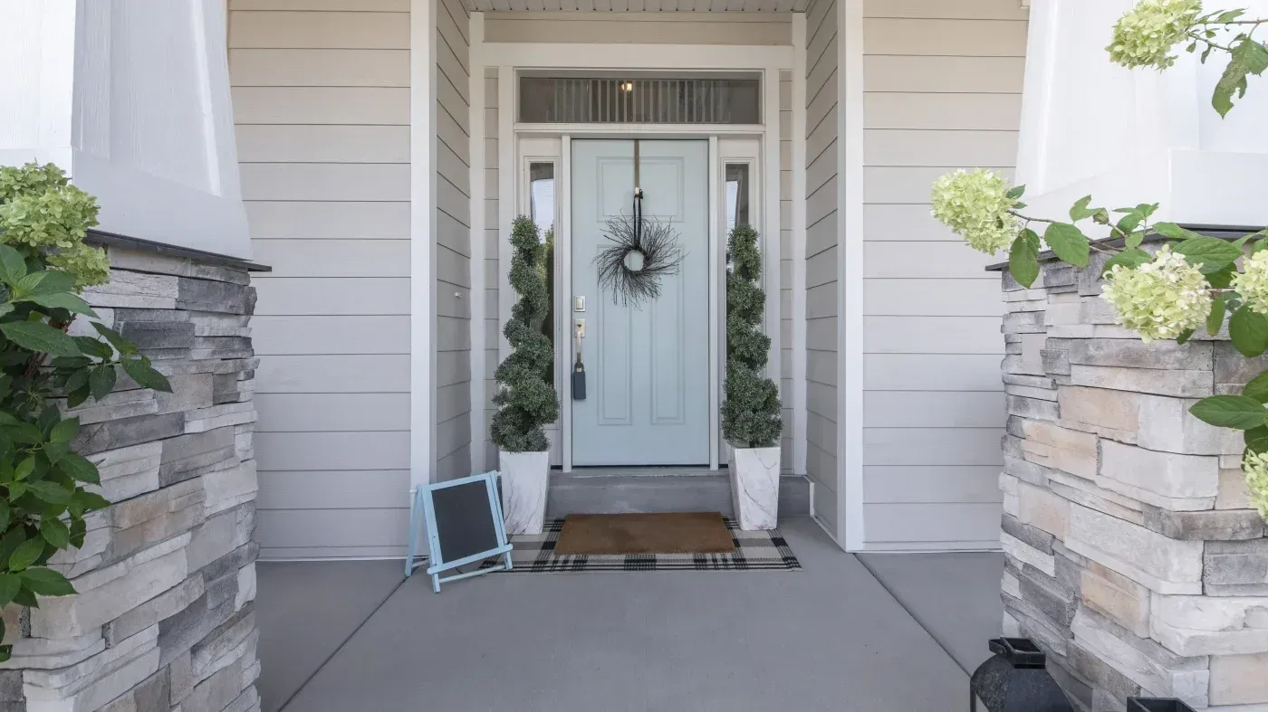 Front porch with light blue double doors, flanked by potted topiaries and stone pillars, decorated with a black wreath and welcome mat