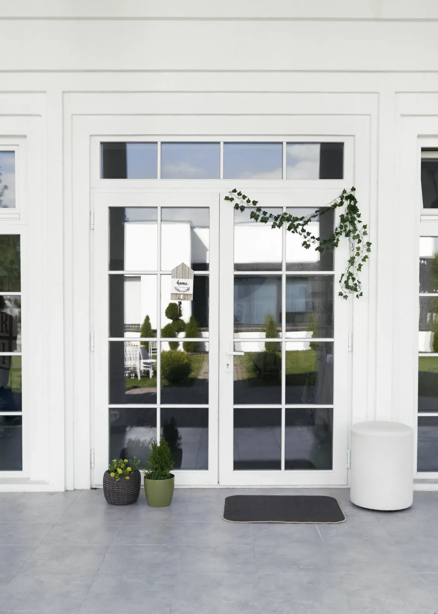 White modern glass double doors with transom window, flanked by potted plants and a cylindrical planter, opening to a patio area
