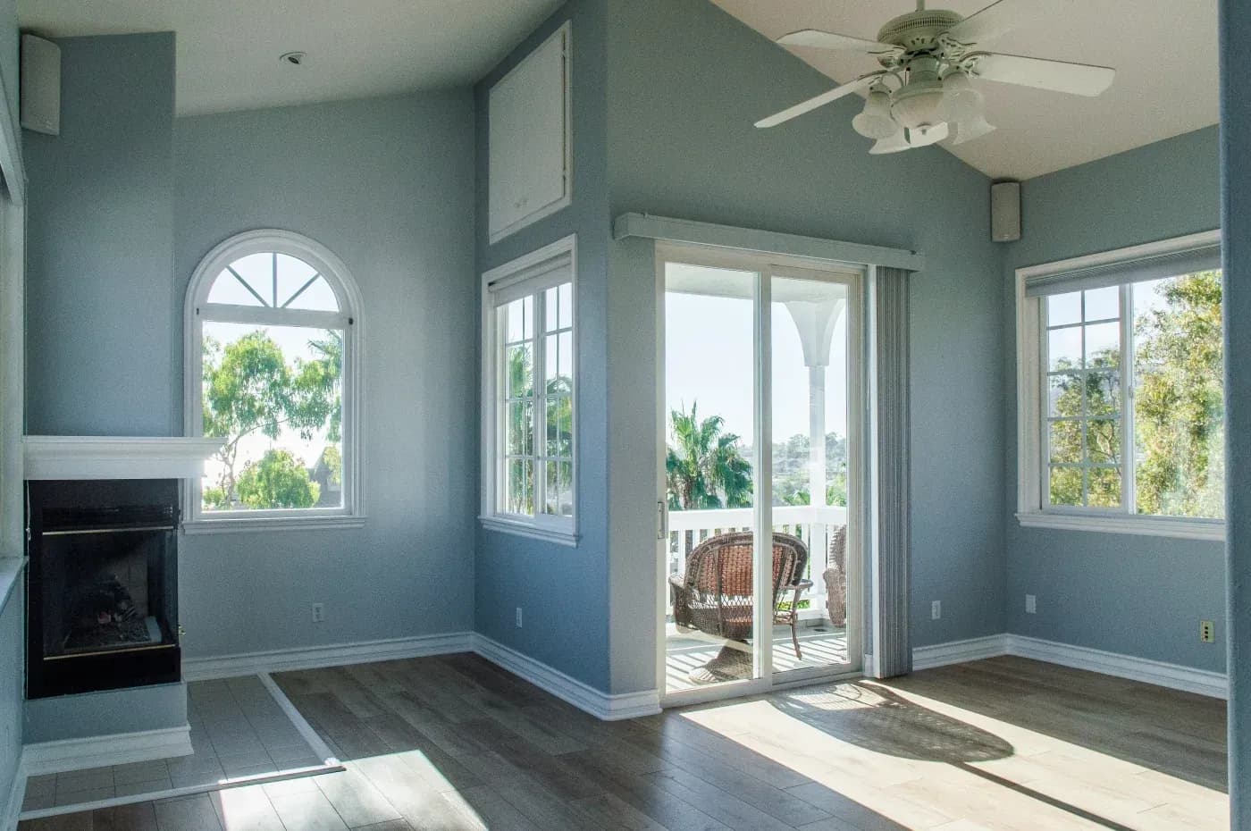 Bright living room with blue-green walls, fireplace, ceiling fan, and multiple windows including French doors opening to a deck with outdoor seating