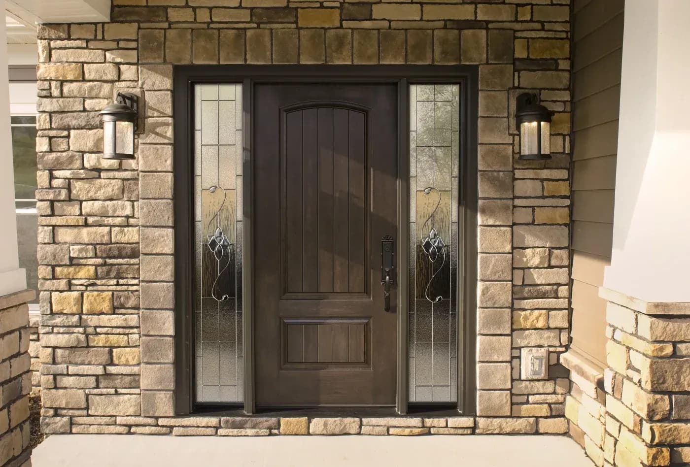 Dark wooden front door with decorative glass sidelights flanked by tan stone brick, wall sconces on either side, and black hardware
