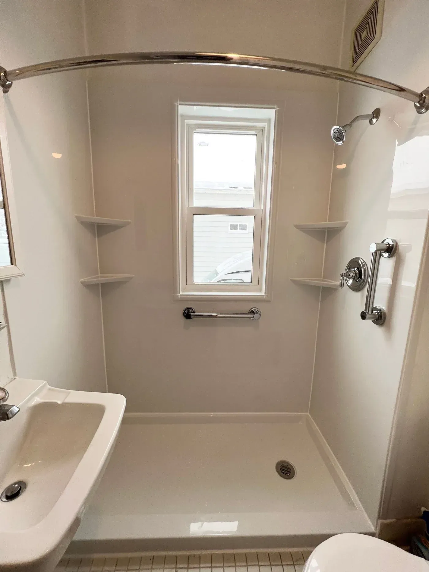 Modern bathroom shower enclosure with beige walls, corner shelves, chrome fixtures, grab bar, showerhead, and window overlooking exterior