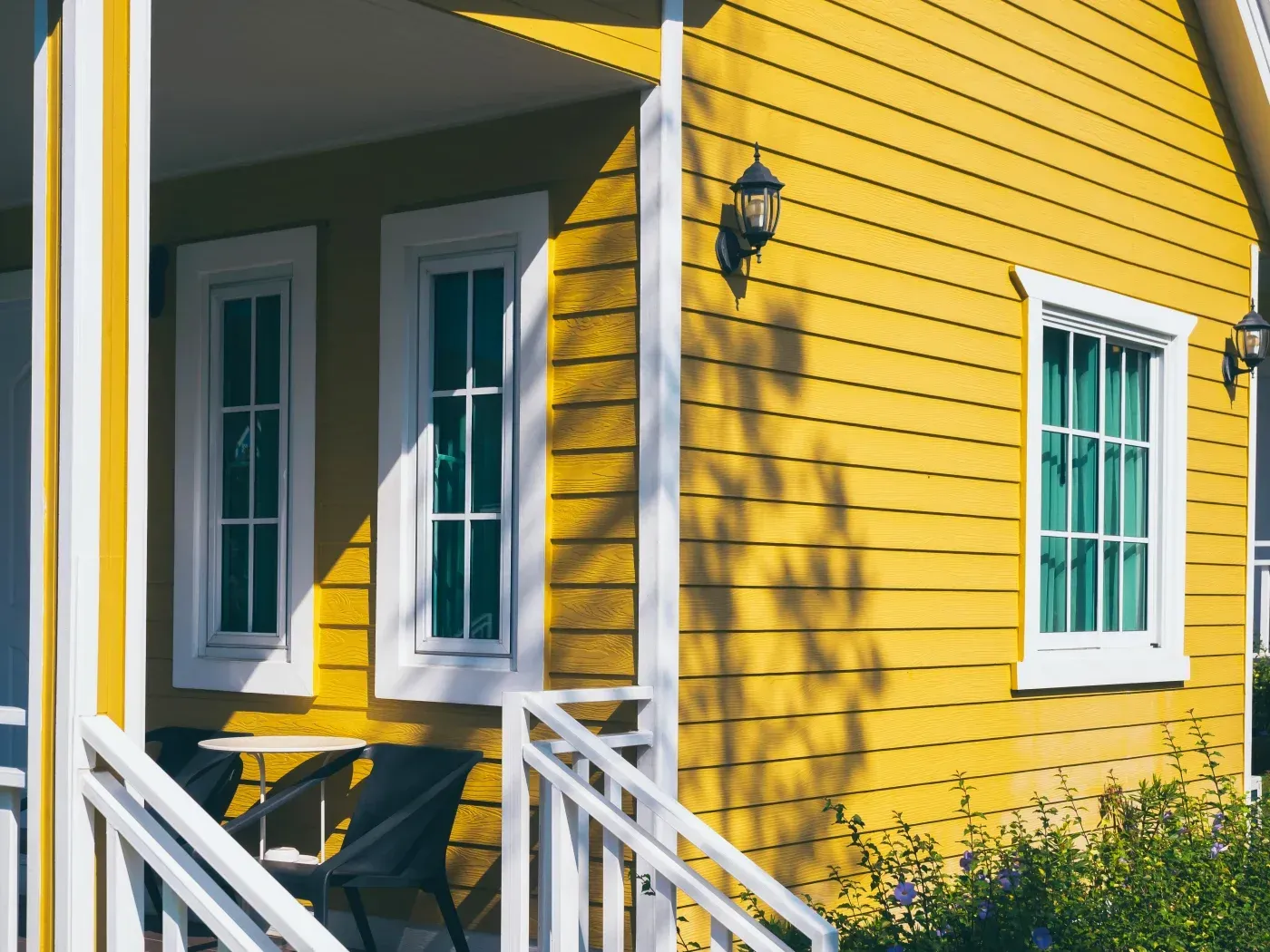 Bright yellow house exterior with white trim, featuring a covered porch, multiple windows with green panes, white columns, and wall-mounted lanterns casting shadows on the siding