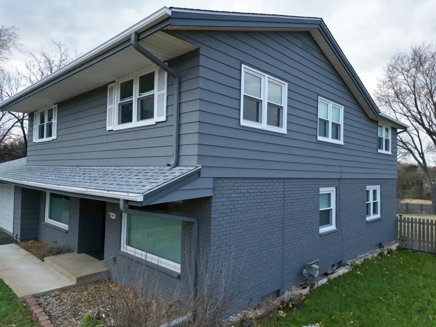 Two-story blue house with white trim windows, brick foundation, attached garage with green door, and front lawn with landscaping