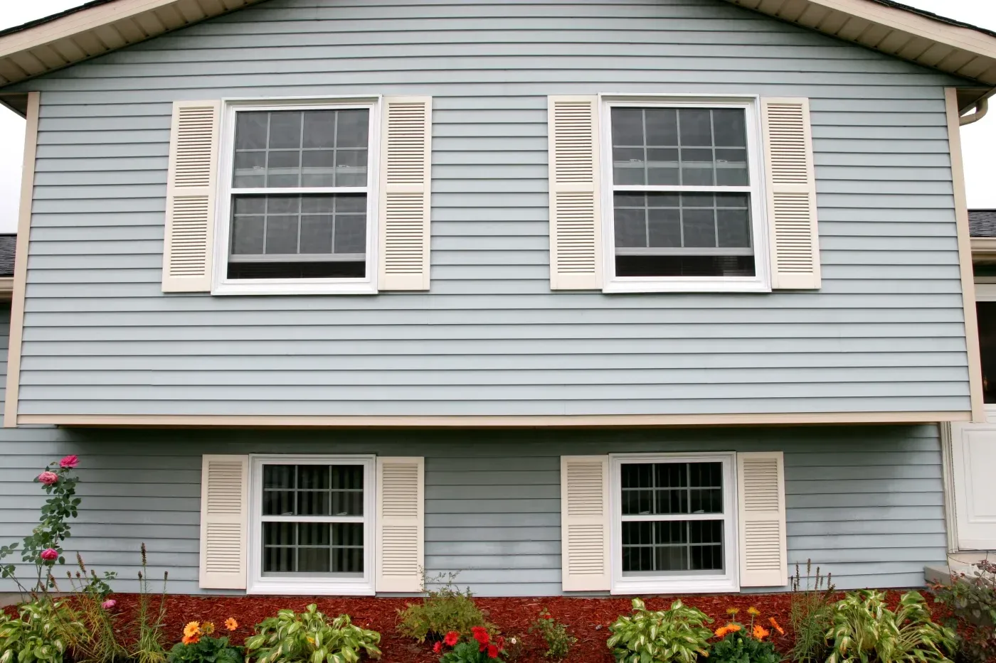 Two-story house with light blue horizontal siding, white trim, tan shutters on four windows, and a flower garden with red mulch and colorful blooms in the foreground