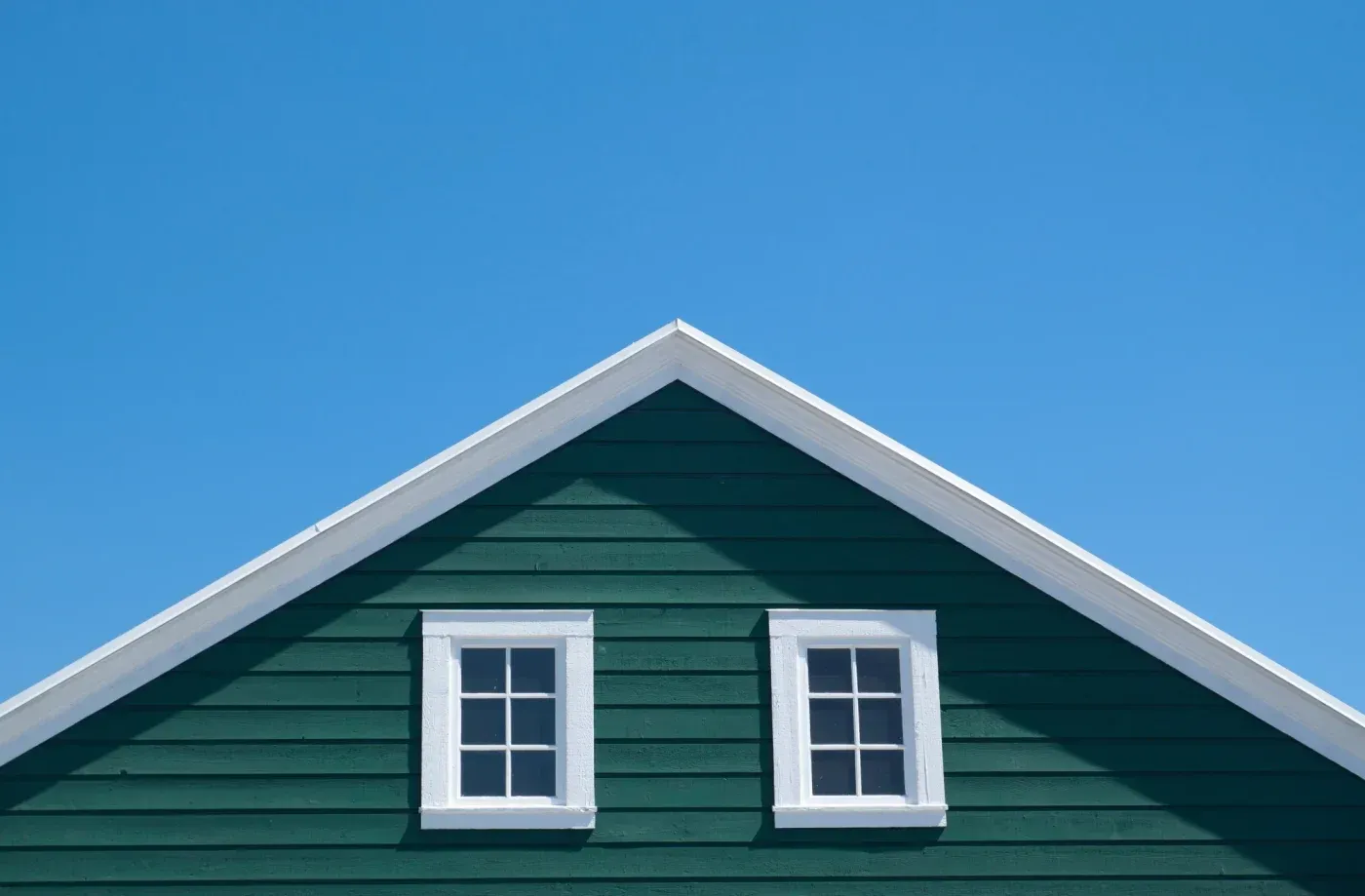Green house gable with white trim and two white-framed windows against clear blue sky