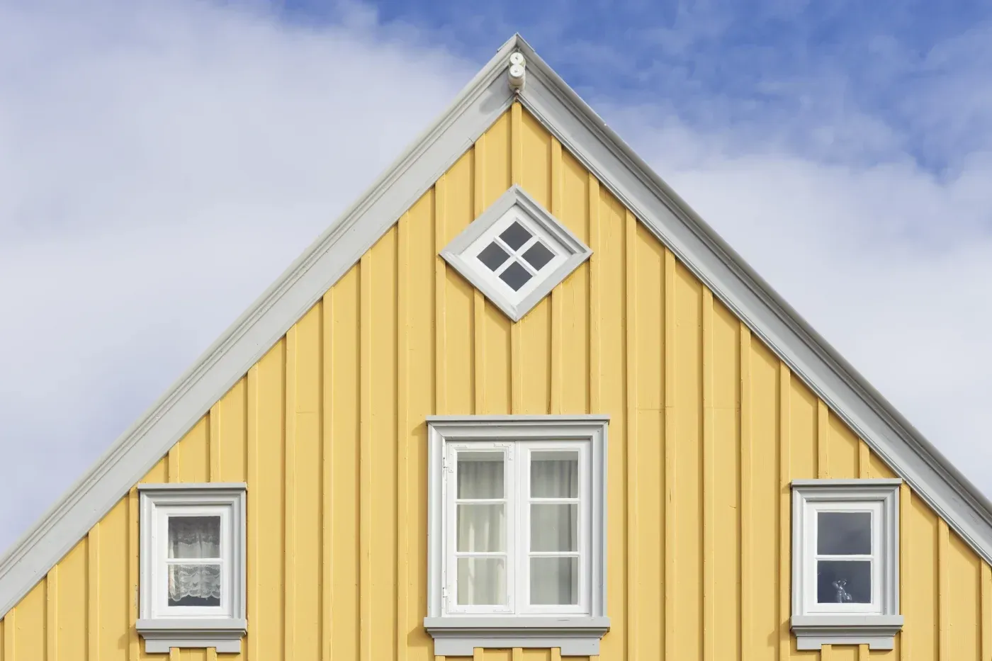 Yellow gabled house with white trim featuring three windows and a small attic window on the pitched roof against a blue sky