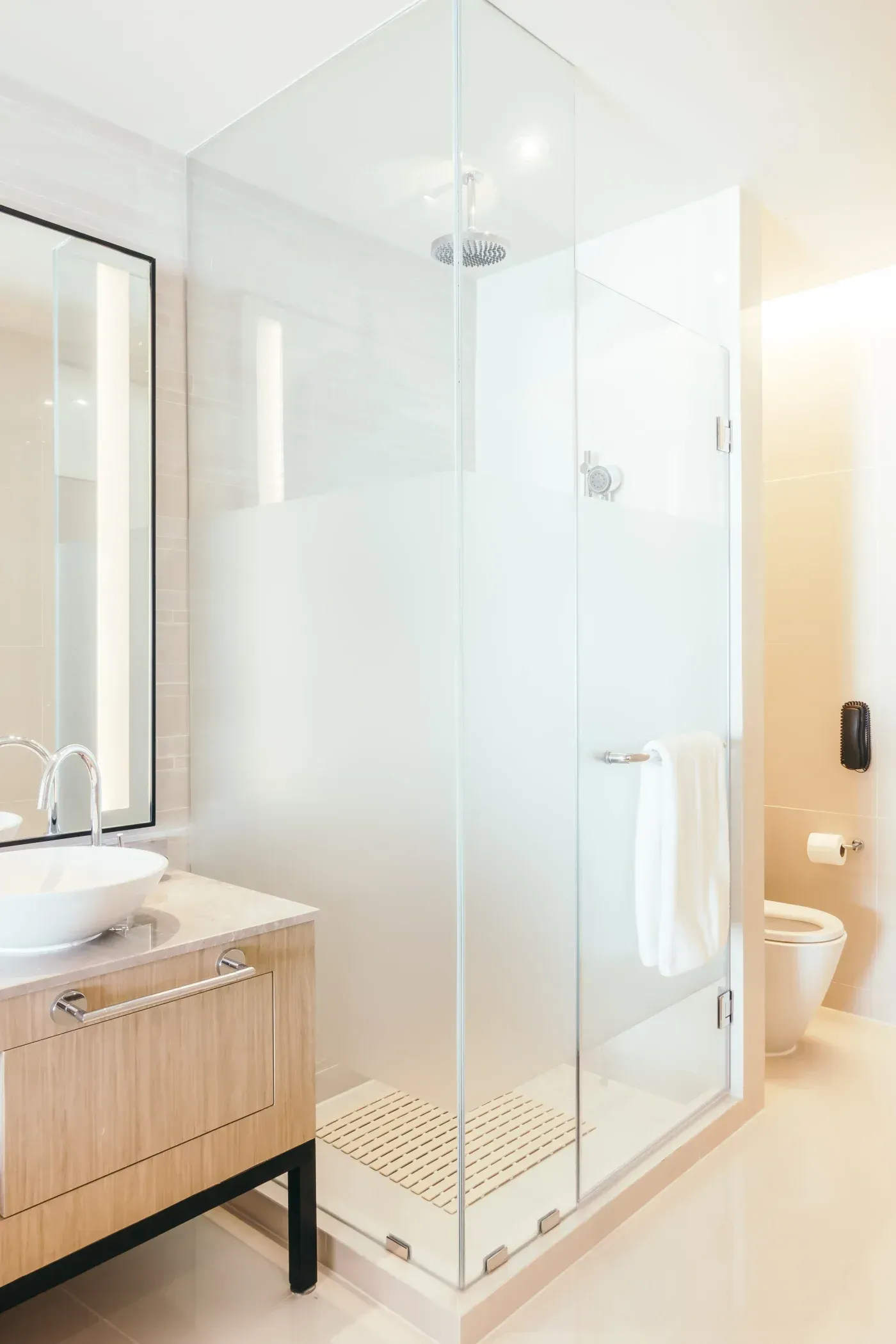 Modern bathroom with frameless glass shower enclosure, wooden vanity with white sink, and toilet, featuring overhead rainfall showerhead and neutral tile finishes