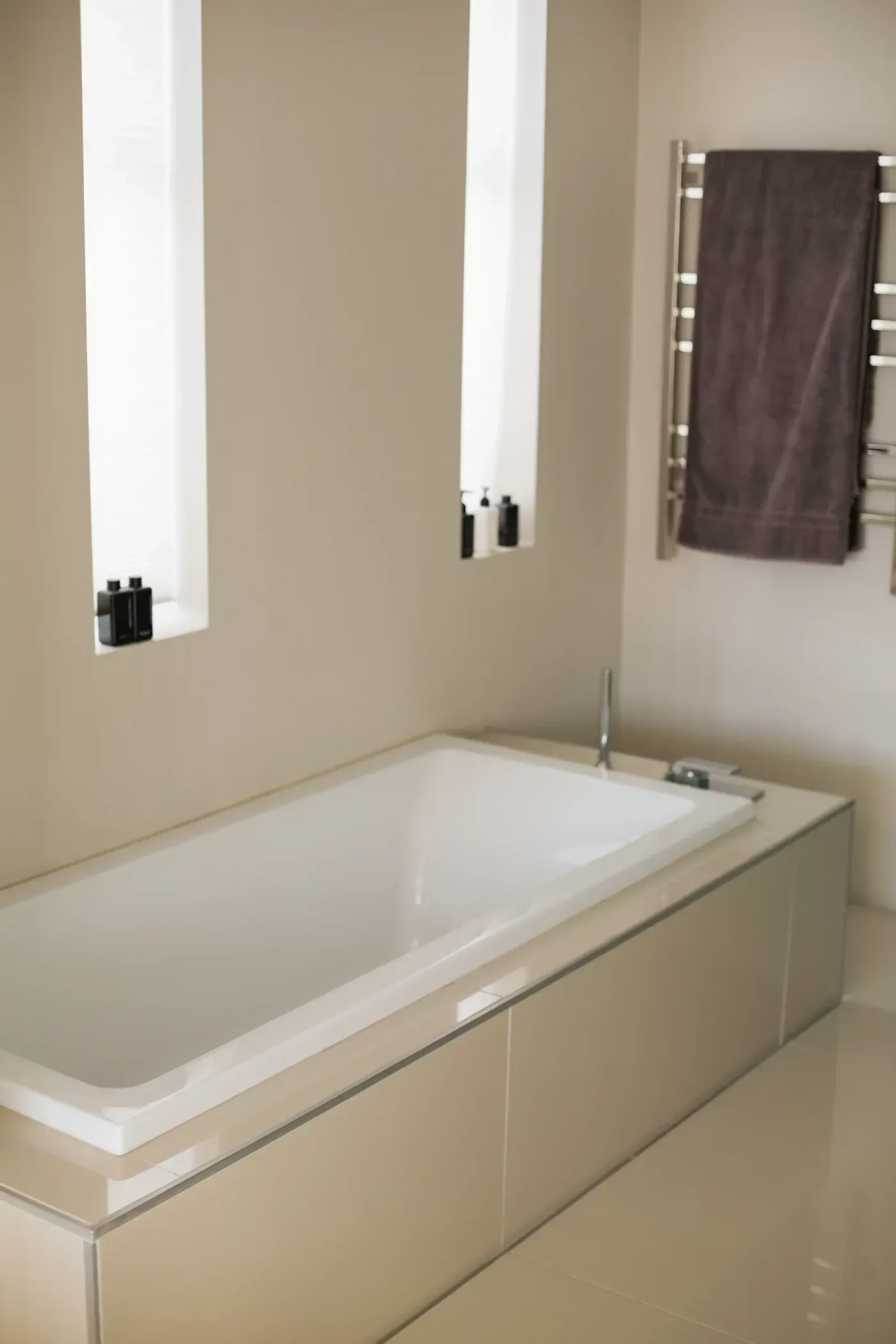 Modern bathroom with white rectangular bathtub, lit mirrors with black canisters, heated towel rack with brown towel, and chrome faucet