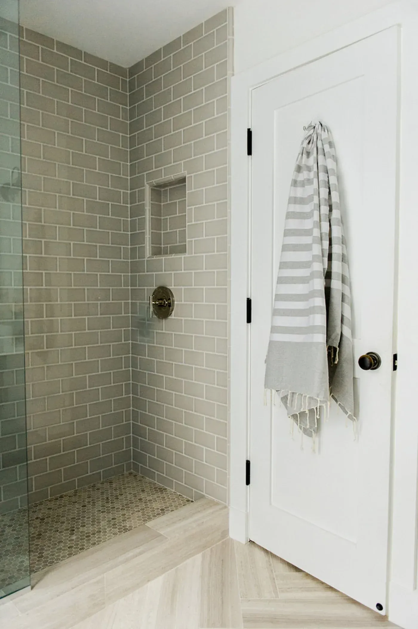 Modern bathroom with glass-enclosed shower featuring beige subway tile walls and mosaic tile floor, white door with black hardware and striped towel hanging outside shower area