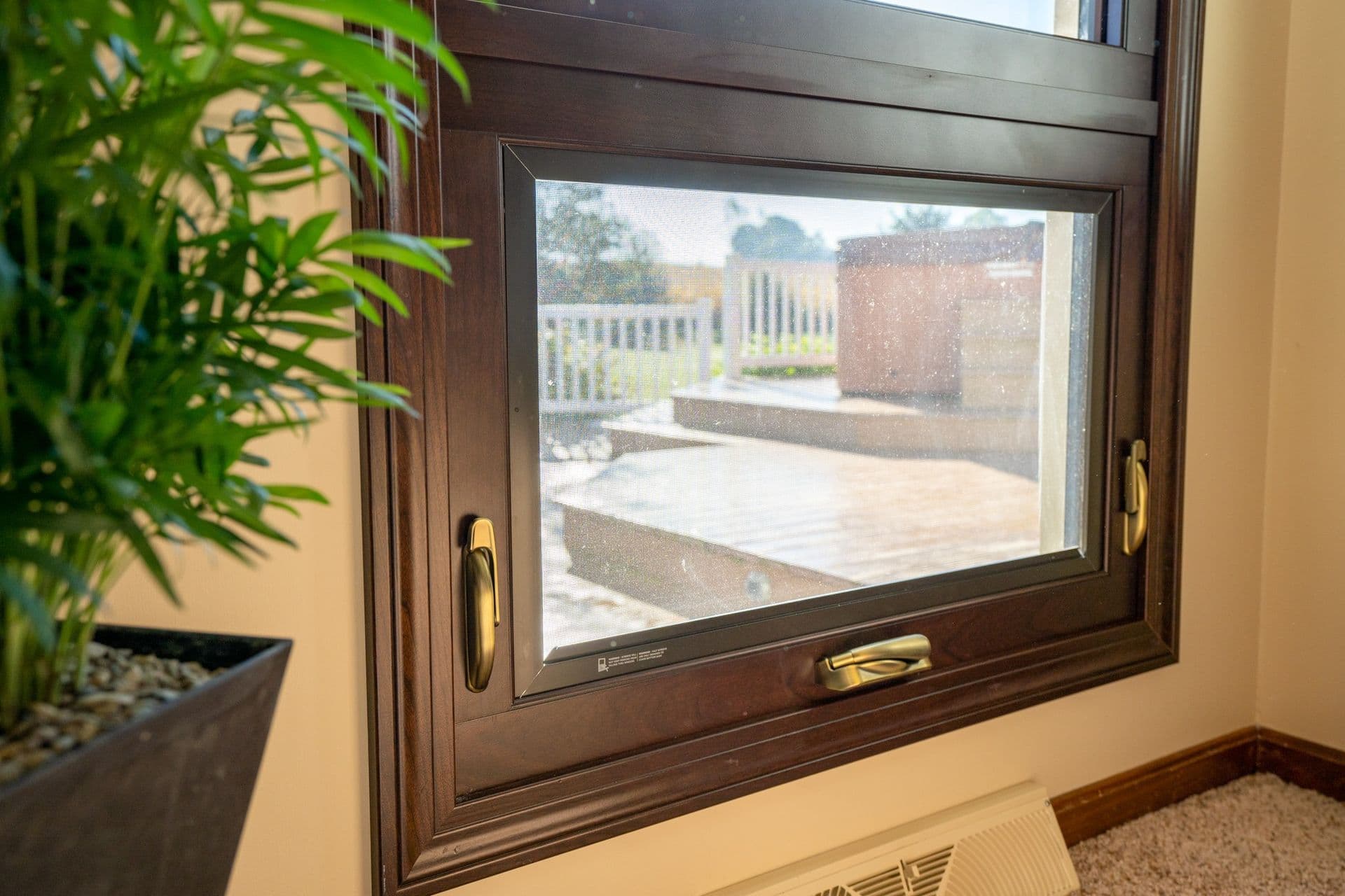 Interior close-up view of an Aeris awning window featuring a rich Hazelnut woodgrain finish, antique brass locks, and a matching fold-down crank handle. The mesh screen provides a view to an outdoor wooden deck and hot tub, set adjacent to a potted indoor plant and floor baseboard vent.