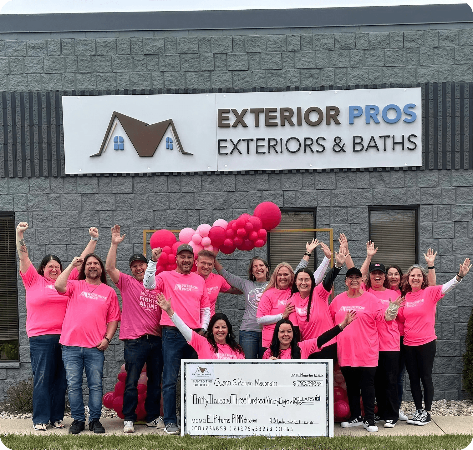 Group photo of the Exterior Pros team standing outside their grey brick building beneath the "EXTERIOR PROS EXTERIORS & BATHS" business sign. Many team members are wearing bright pink shirts, celebrating a charitable donation. In the foreground, two women hold a giant presentation check made payable to Susan G. Komen Wisconsin for the amount of $30,398.84, with the memo reading "E.P. turns PINK donation".