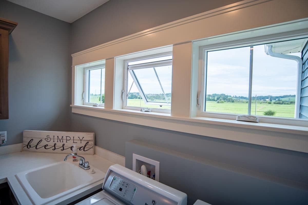 Interior laundry room view featuring a row of three white-framed Endure awning windows set in thick white craftsman-style trim against grey walls. The center window sash is cranked outward to let in ventilation, overlooking an open green rural landscape. A laundry sink and washing machine sit in the foreground next to a decorative sign reading "SIMPLY blessed."