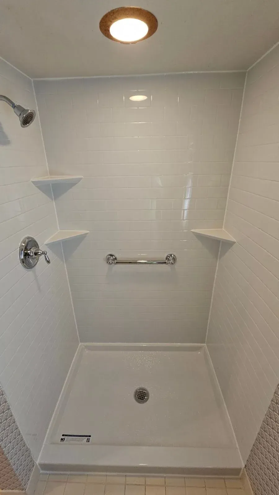 Interior view of a modern walk-in shower stall featuring glossy white subway tile-patterned wall panels, a low-threshold white shower base, chrome hardware including a showerhead and single-handle faucet, a horizontal chrome safety grab bar on the back wall, three integrated white corner shelves, and an overhead recessed ceiling light fixture.