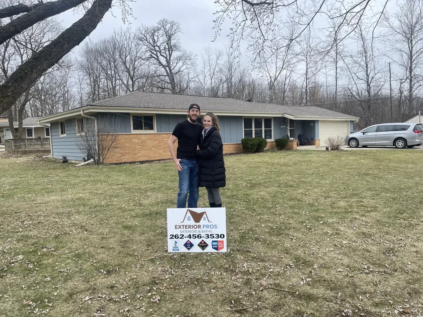 Exterior view of a smiling couple standing in front of a single-story ranch home with blue siding, tan brick accents, and a yard sign for "Exterior Pros Exteriors & Baths."
