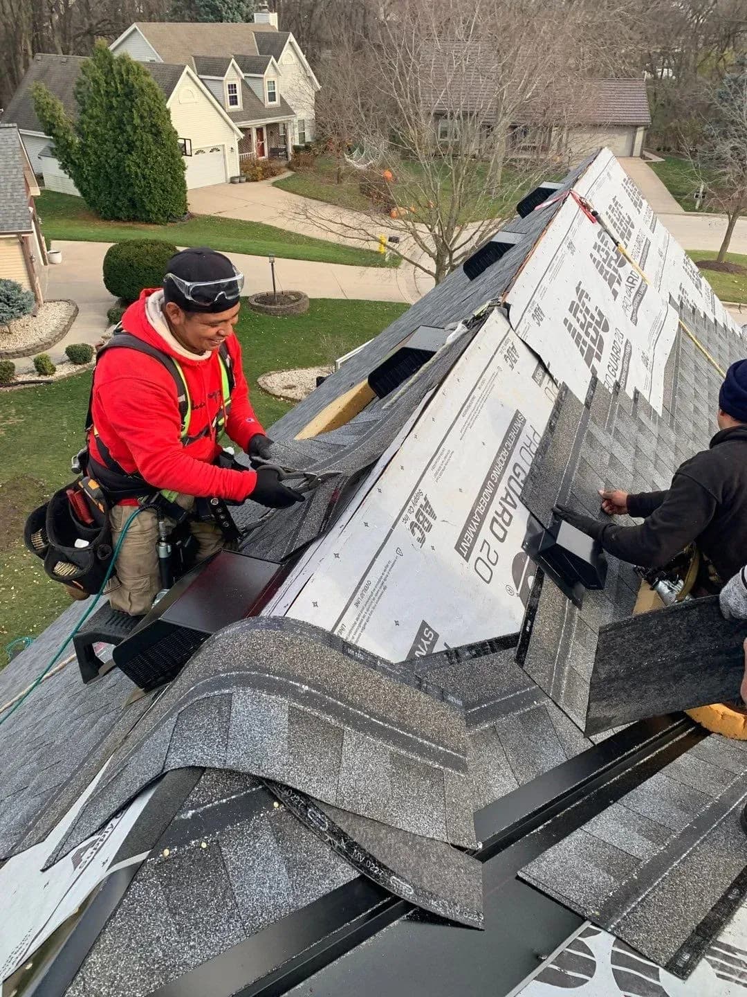 Action shot of roofers wearing safety harnesses installing dark grey asphalt shingles and ridge vents over PRO GUARD 20 synthetic underlayment on a steep residential roof peak.