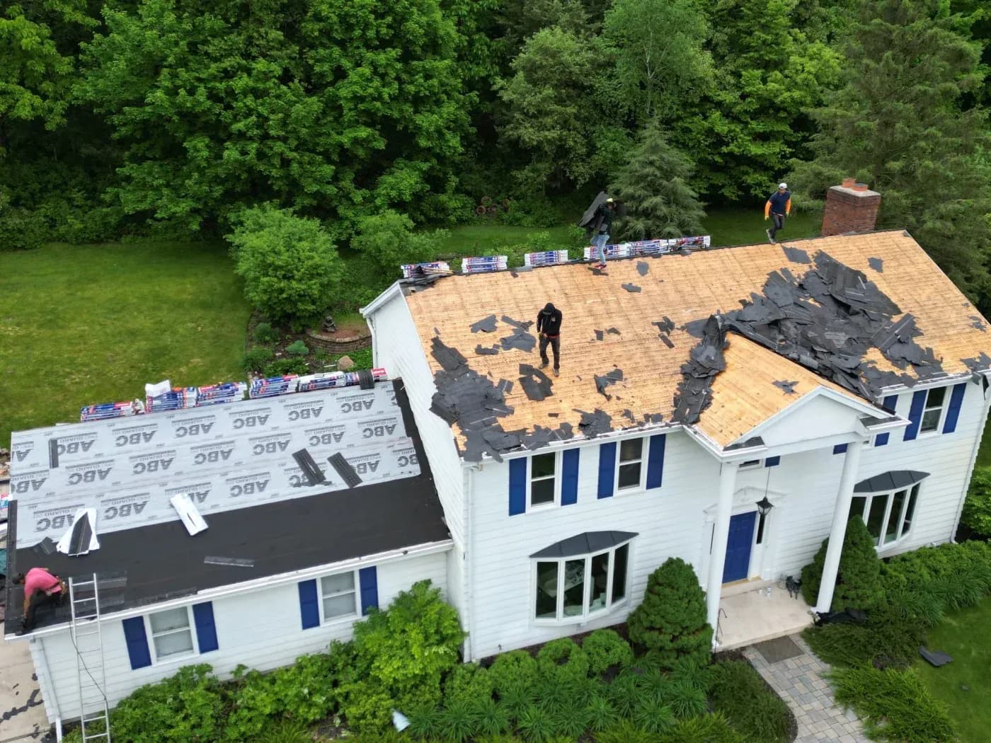 High-angle aerial view of a roofing crew tearing off old asphalt shingles down to the wooden decking on a two-story white home with blue shutters, while preparing adjacent roof sections with synthetic underlayment and new shingle bundles.