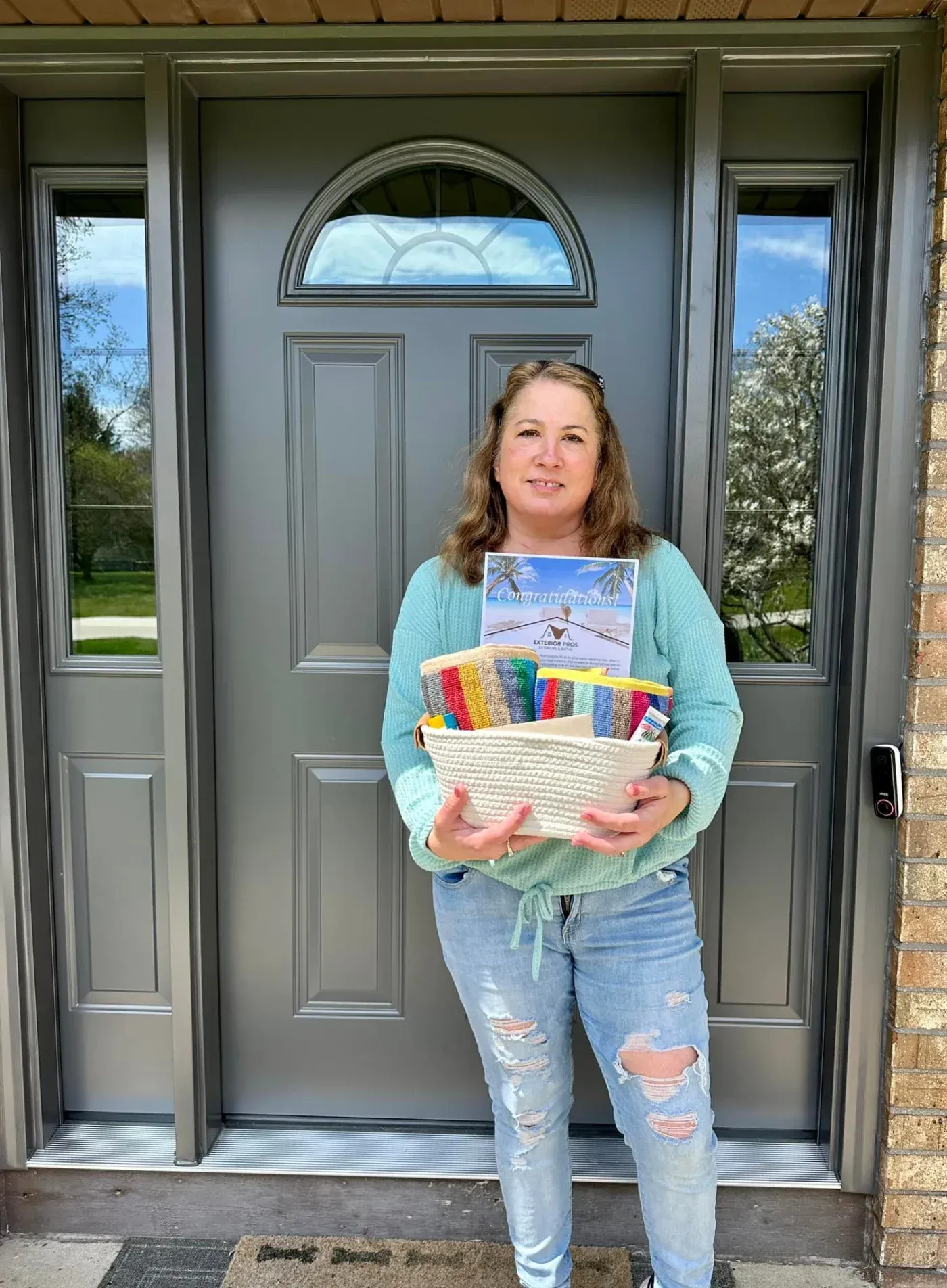 A woman standing in front of a newly installed charcoal grey front door with glass sidelights, holding a woven gift basket and a "Congratulations!" card from Exterior Pros.