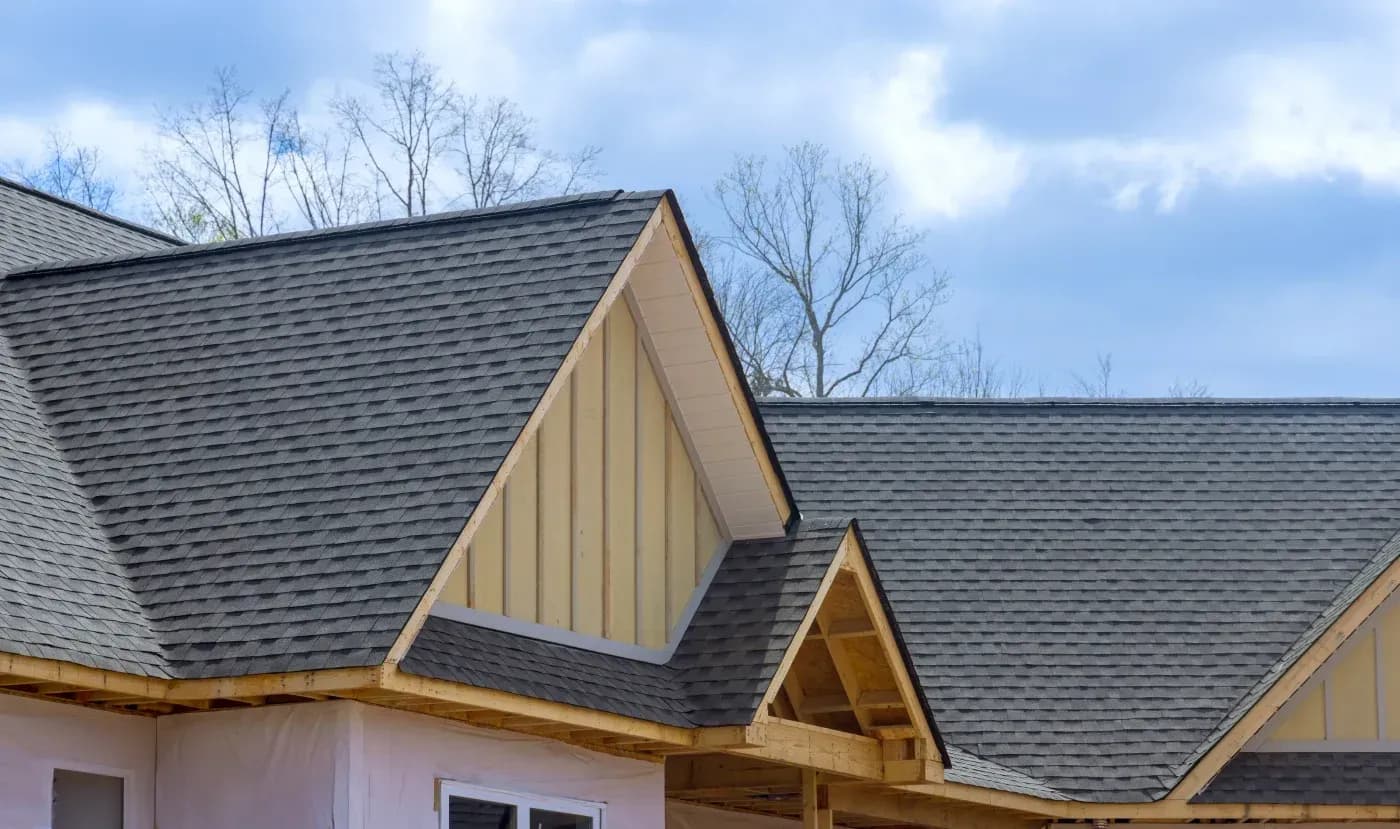 Exterior view of a multi-gabled home roof under construction featuring new dark grey asphalt shingles, yellow board-and-batten accent siding within the upper gable peaks, exposed wooden fascia and framing, and house wrap covering the lower exterior walls under a cloudy blue sky.