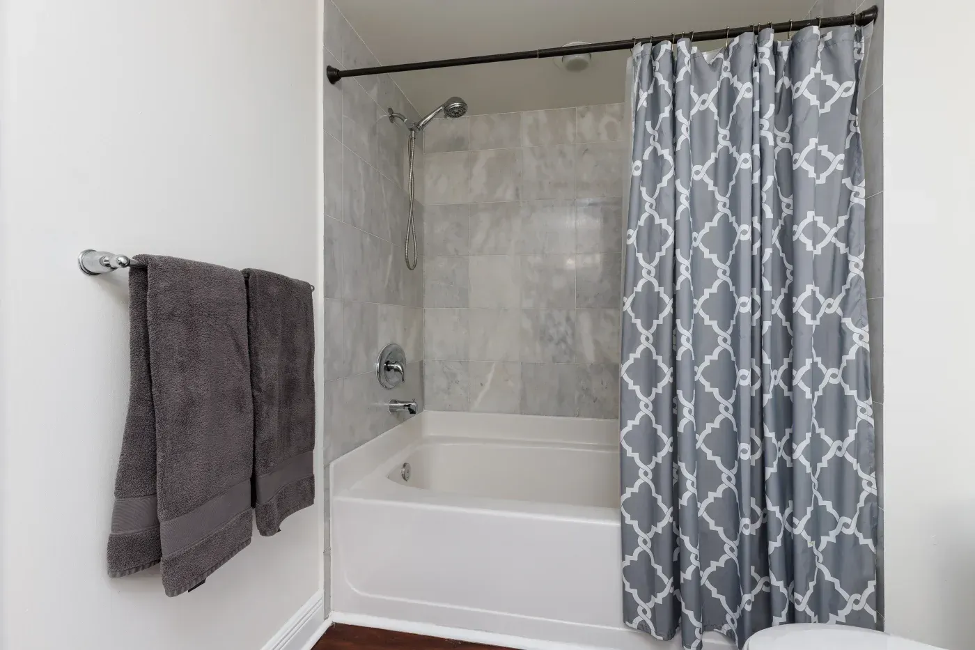 Bathroom with white bathtub, gray tile surround, gray geometric patterned shower curtain on black rod, and gray towels on wall-mounted rack