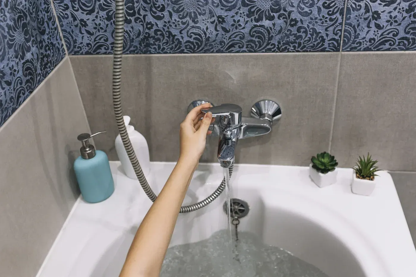 Person adjusting a handheld shower head over a bathtub with soap dispenser and small potted plants on the tub edge