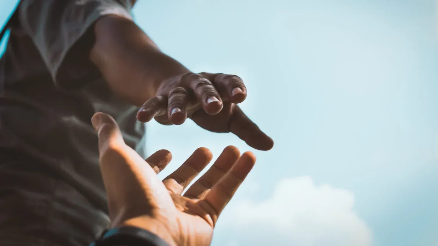 Two people reaching toward each other with outstretched hands against a bright blue sky.