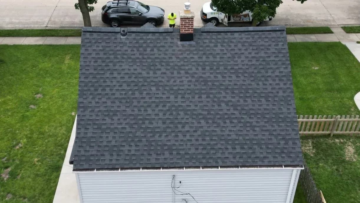 Aerial view of a gray asphalt shingle roof with a worker in a safety vest standing on top near a brick chimney