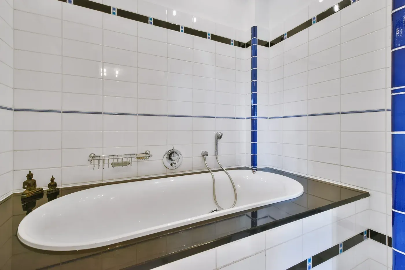Modern bathroom with white subway tile walls, blue accent tiles, and oval bathtub with chrome fixtures and hand-held shower head on dark countertop