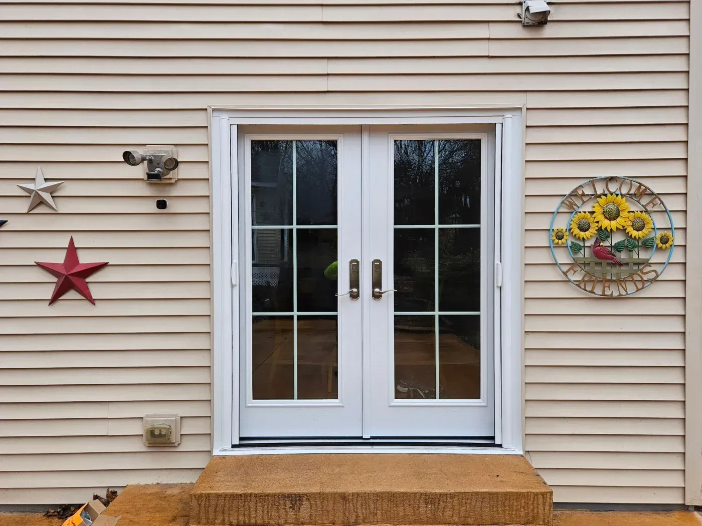 White double glass patio doors with motion sensor light above, flanked by red and silver decorative stars and a sunflower wall medallion on tan siding