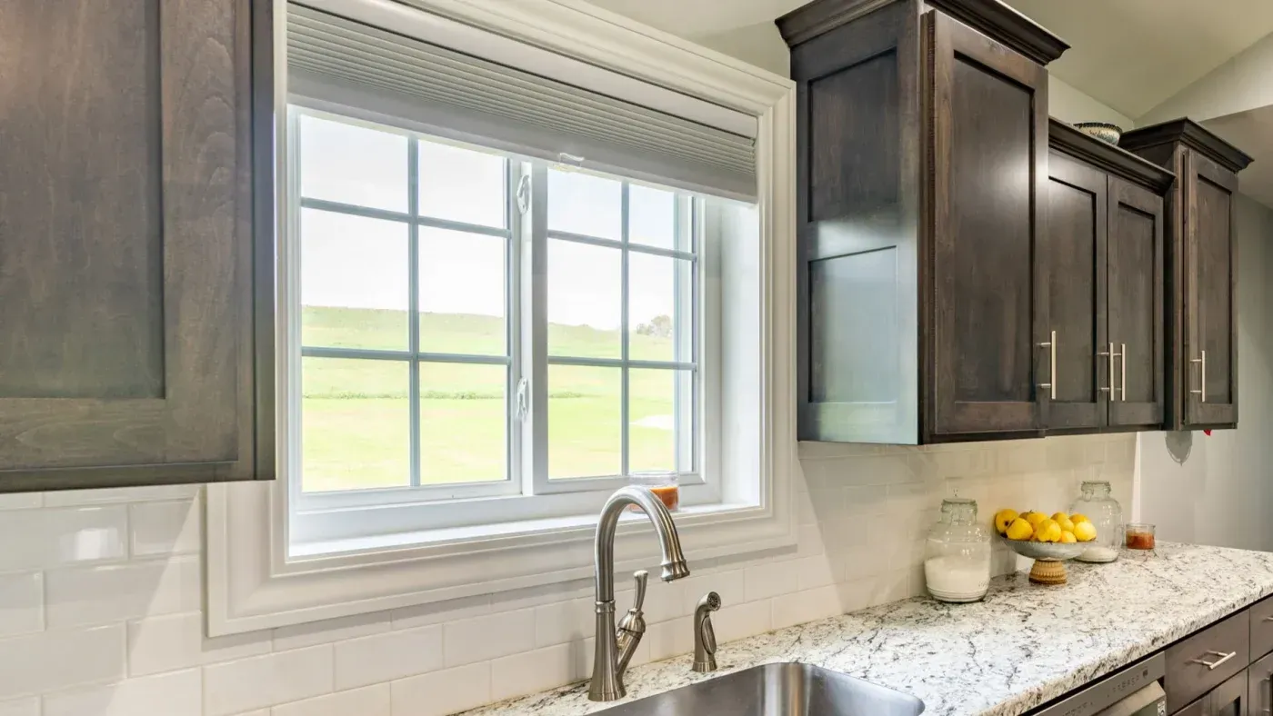 Modern kitchen with stainless steel sink beneath large window overlooking green fields, granite countertop, white subway tile backsplash, and dark wood cabinetry with glass doors