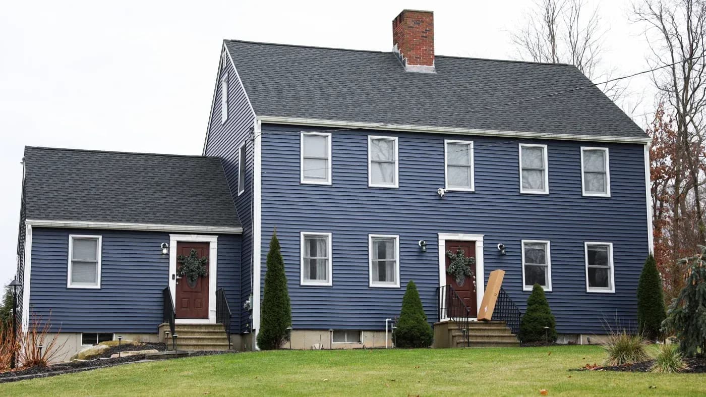 Two-story blue colonial house with brick chimney, white trim windows, red doors, and attached single-story wing on the left
