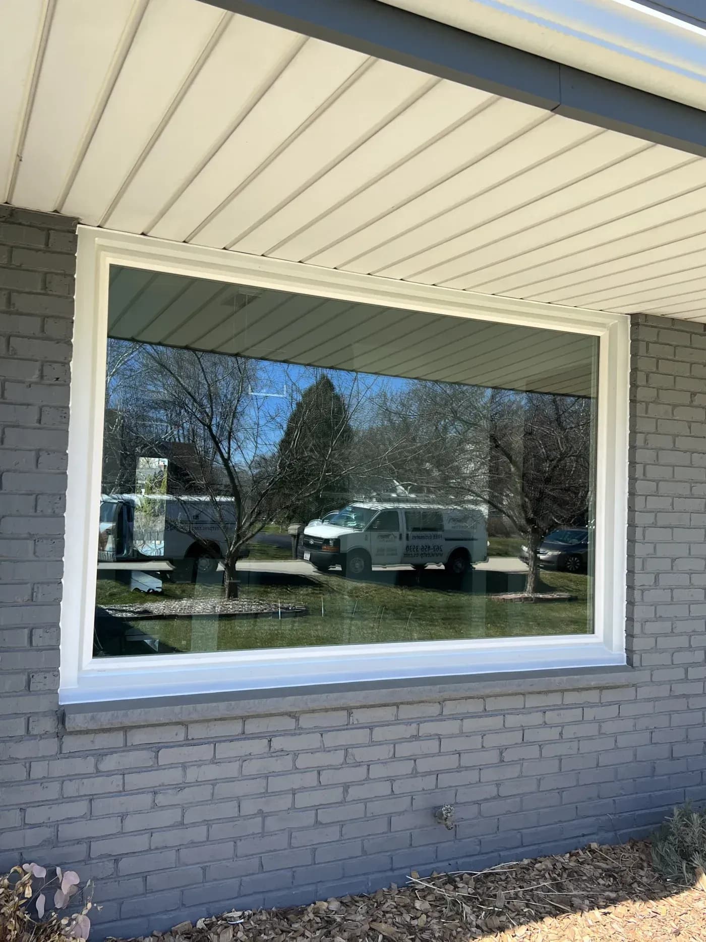 Large picture window with white frame installed in brick wall under retractable pergola awning, showing driveway with service van and bare trees