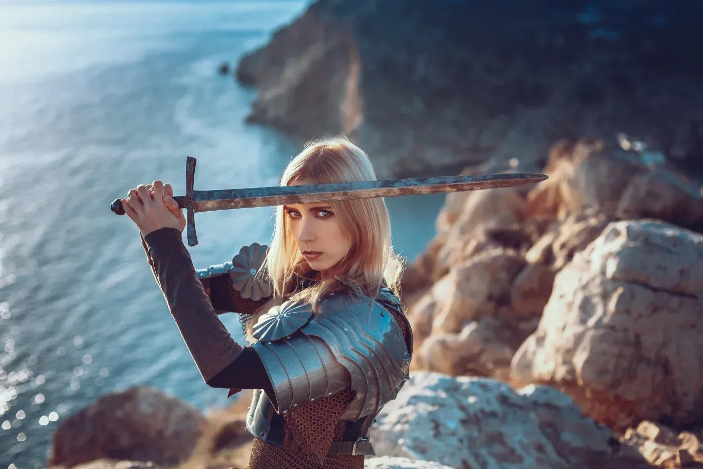 Woman in medieval-style armor holding a sword across her face on a rocky coastline.