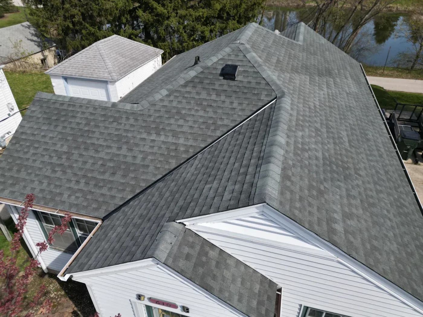 Aerial view of residential house with gray asphalt shingle roof, metal standing seam roof sections, attached garage, and white cupola, surrounded by trees and water