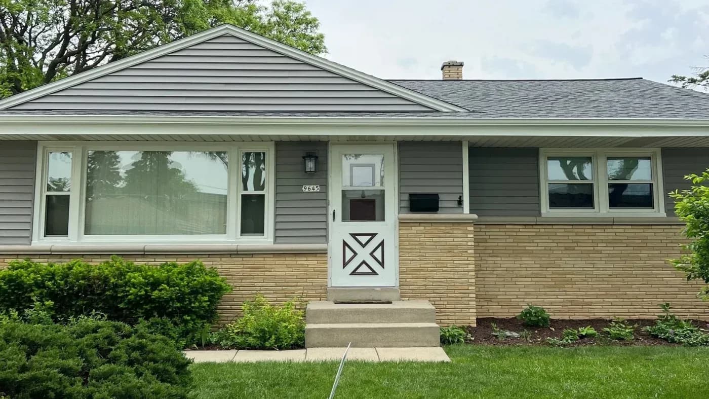 Exterior of single-story ranch house with tan brick and gray siding, white-trimmed windows, white entry door with X pattern, address 9645, pitched roof, and landscaped front yard