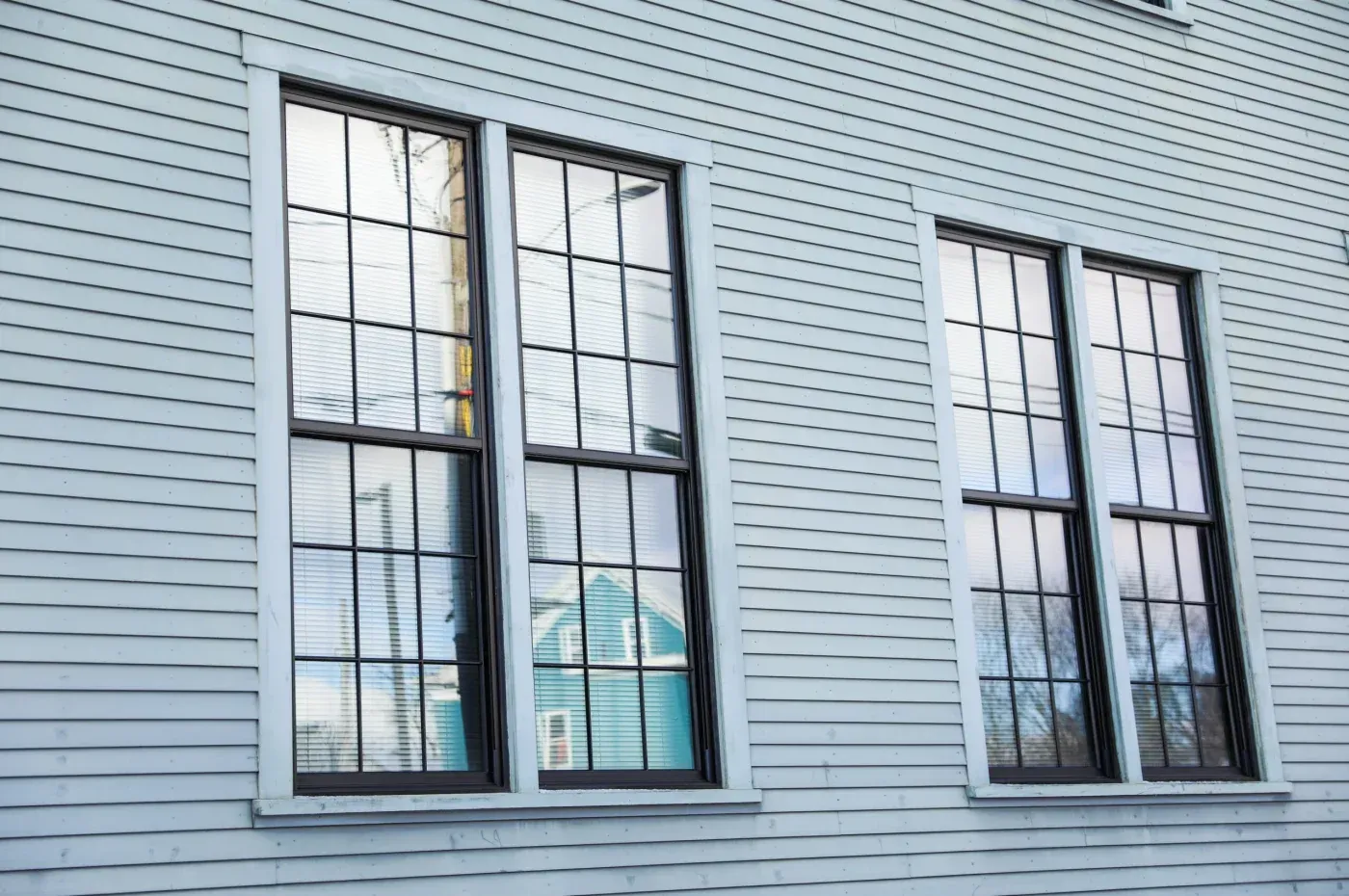 Exterior view of four tall black-framed replacement windows with grid patterns installed on a light-colored siding wall.