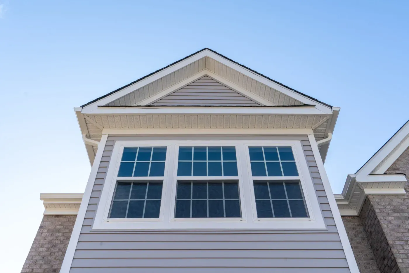 Low-angle exterior view of three white-framed replacement windows with grid patterns beneath a gabled roof and light siding.