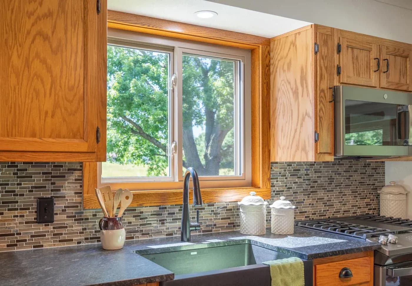 Kitchen with wood cabinetry, dark countertops, brick backsplash, and window overlooking large tree