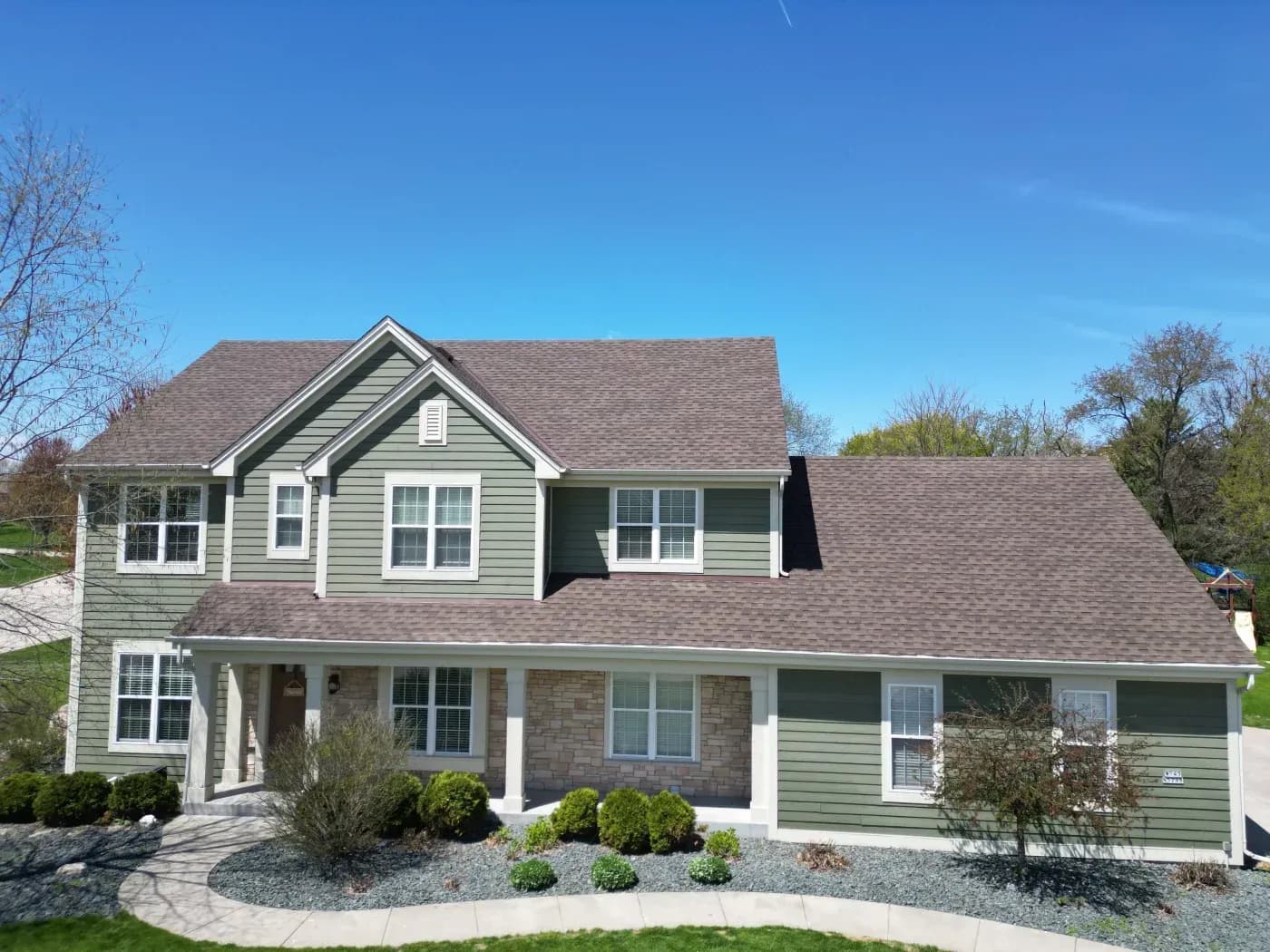 Two-story residential house with green and white siding, stone accents, white trim, and brown roof, featuring a covered front porch and landscaped front yard with shrubs