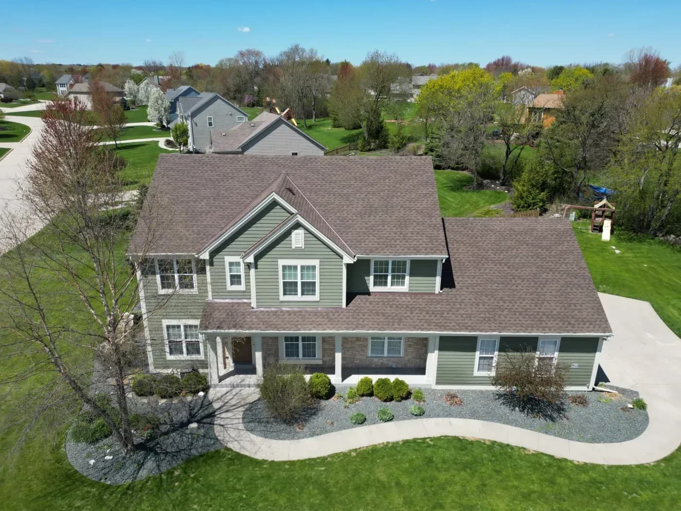 Aerial view of a two-story green home with a large brown asphalt shingle roof, attached garage, landscaped yard, and surrounding neighborhood.