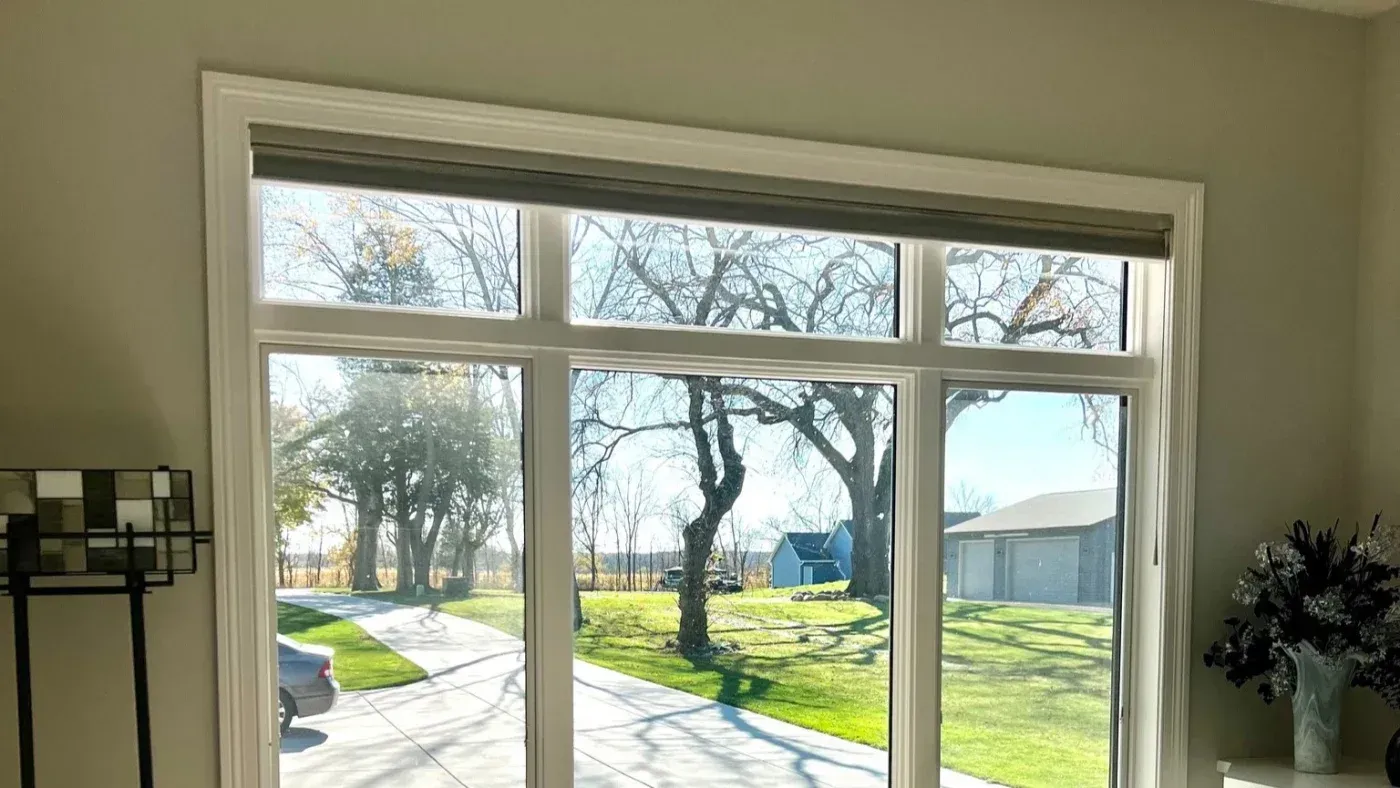 Large sliding glass doors with transom windows overlooking a rural property with mature trees, manicured lawn, driveway, and outbuildings