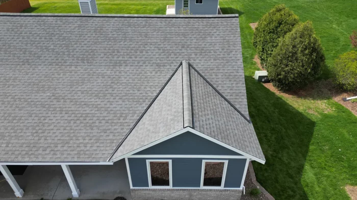 Aerial view of a residential home with gray shingle roof, peaked metal roof cupola, and dark blue siding with white trim windows, surrounded by green lawn and evergreen shrubs
