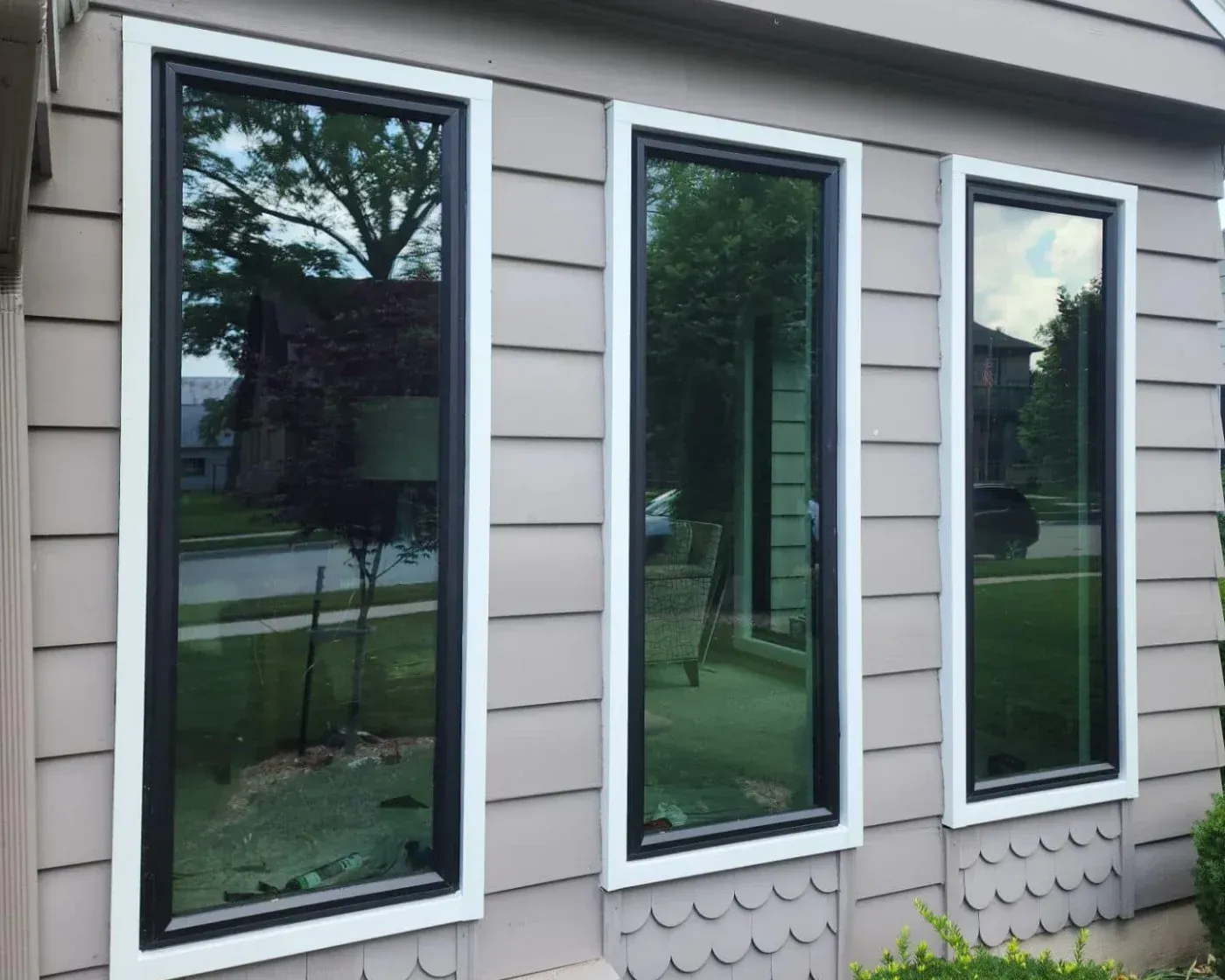 Three tall black-framed windows with white trim on gray siding, reflecting trees and landscape view of residential property