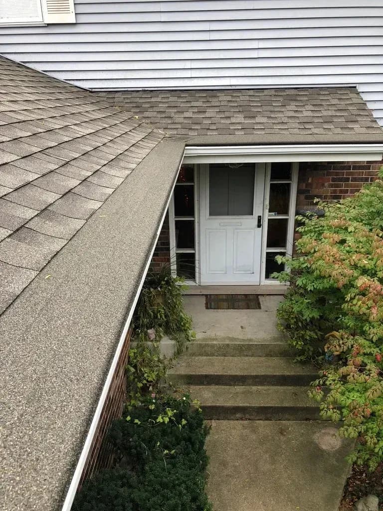 House exterior showing shingled roof, white siding, and patio door entrance with concrete walkway surrounded by climbing ivy and vegetation