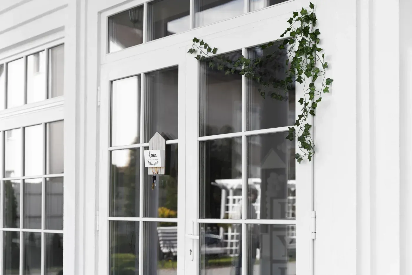 Modern white building facade with glass doors and windows, featuring a small home decoration sign and climbing ivy plant on the right side