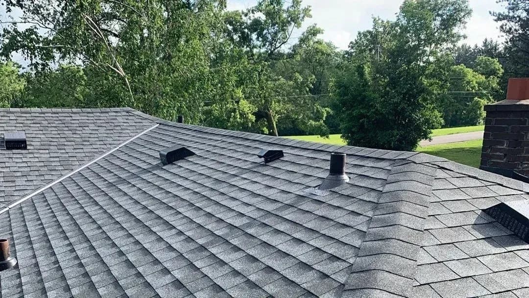 Gray asphalt shingle roof with ridge caps, roof vents, plumbing vents, and a brick chimney on a residential home.