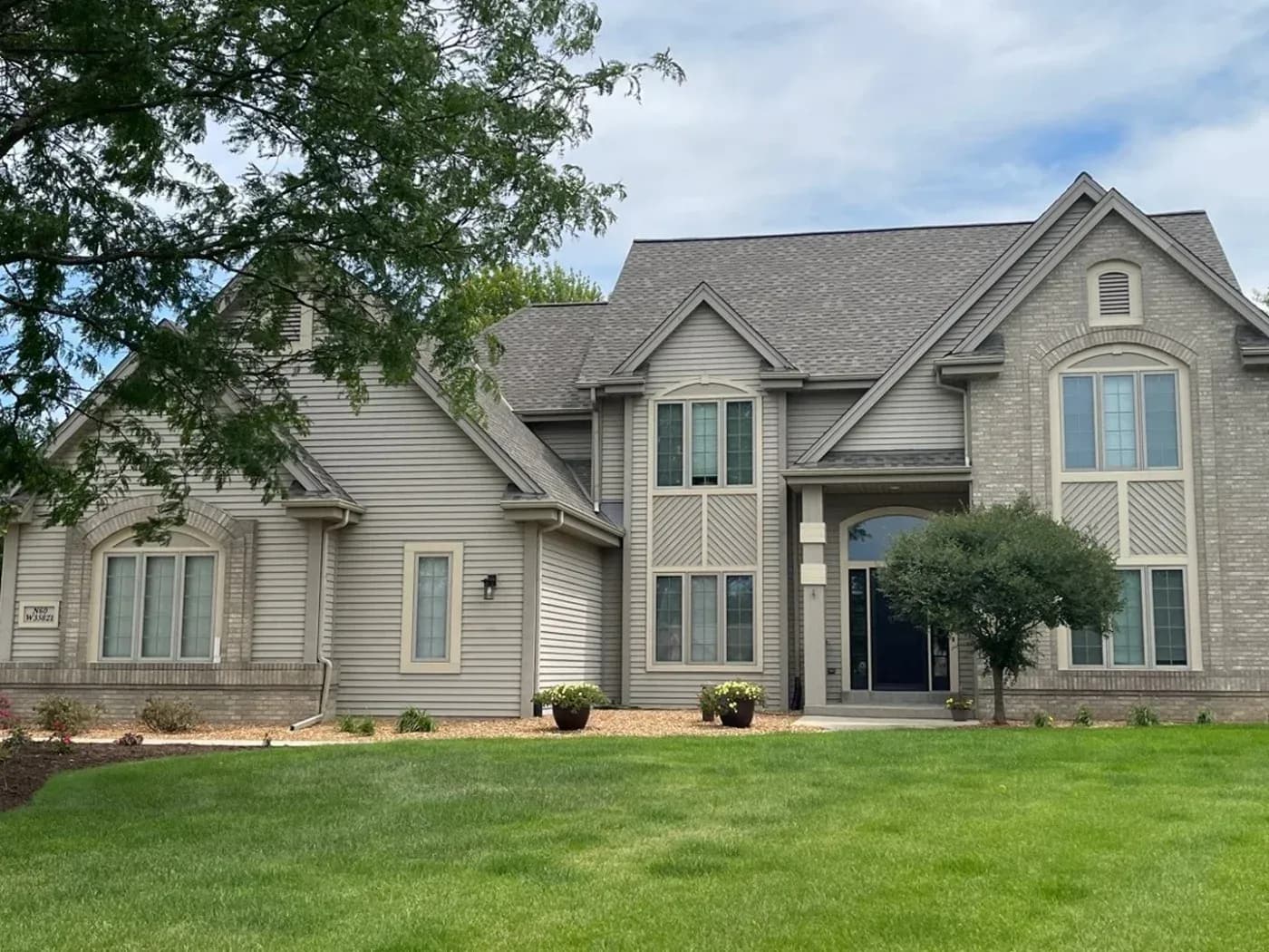 Large two-story home with a gray asphalt shingle roof, beige siding, brick accents, and manicured lawn in Adell, Wisconsin.